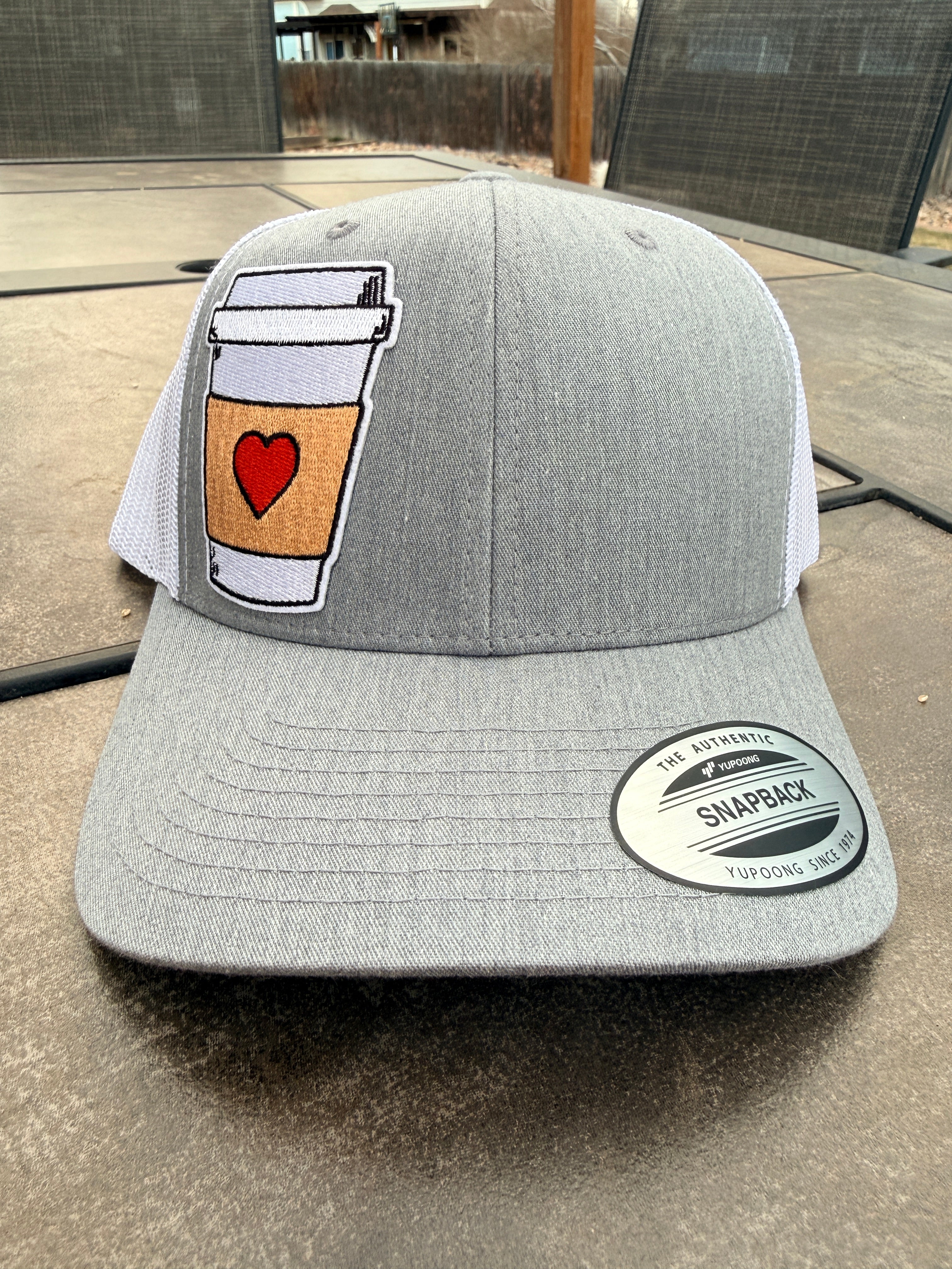 Grey Yupoong snapback trucker hat flat lay with coffee cup heart patch – personalized custom hat – Hedrick Hat Co